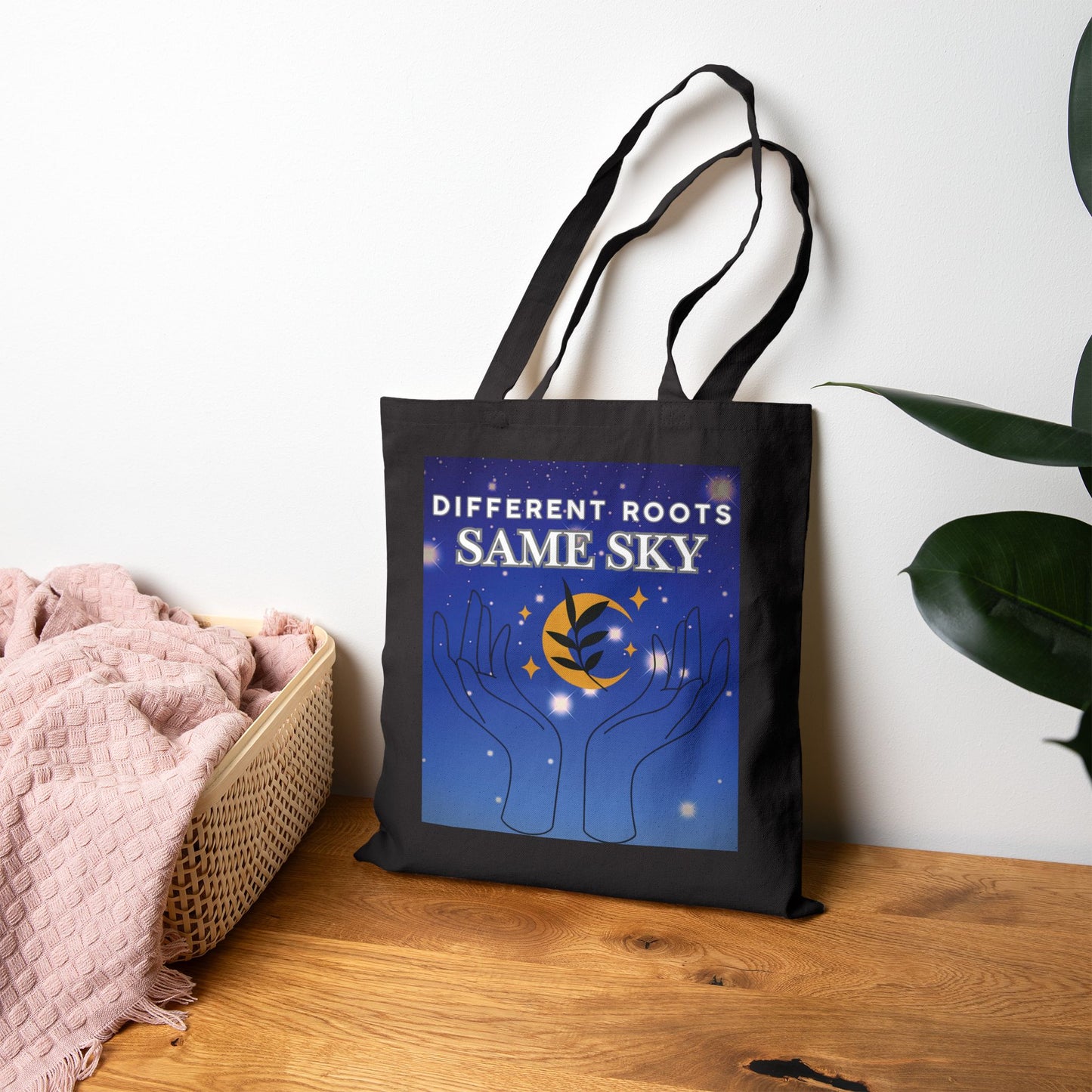 Black tote bag with 'Different Roots Same Sky' design on a wooden surface with a plant in the background