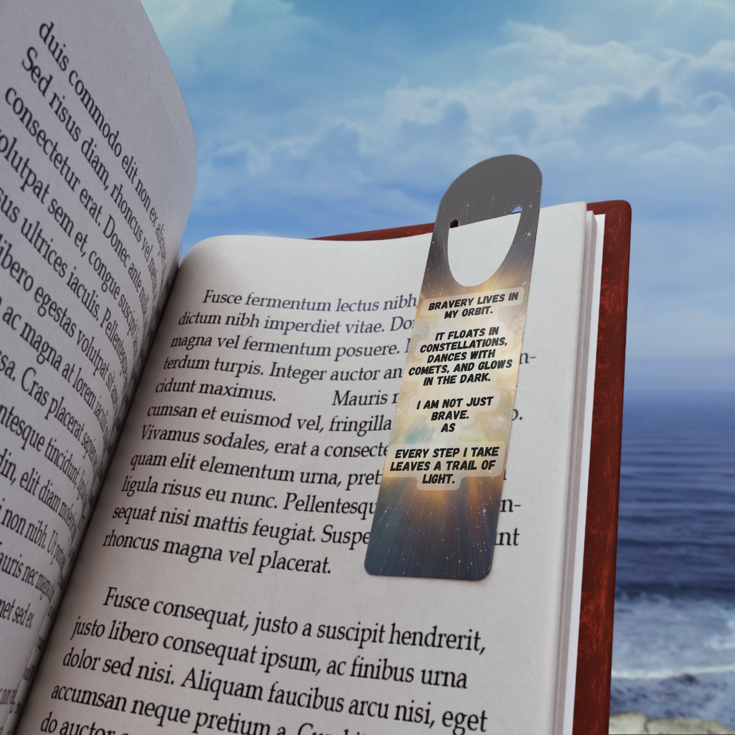 Open book with a metallic bookmark featuringBravery Lives In My Orbit , against a scenic background of water and sky.
