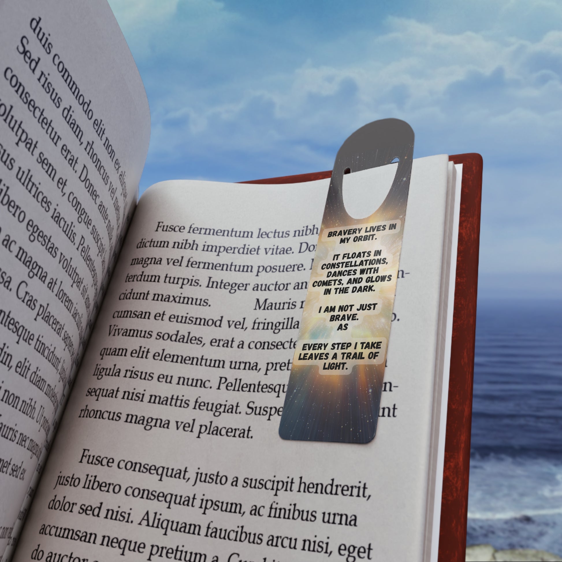 Open book with a metallic bookmark featuringBravery Lives In My Orbit , against a scenic background of water and sky.