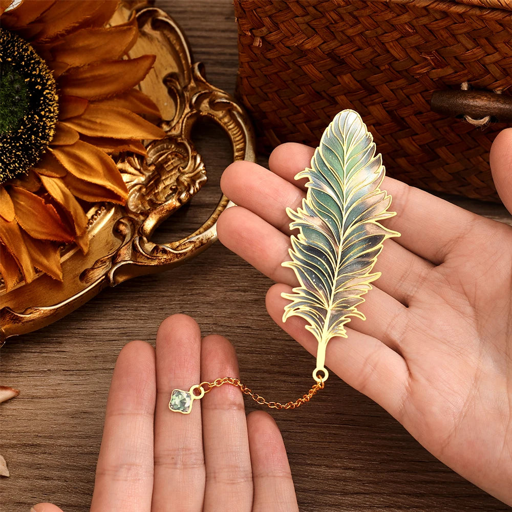 Gold feather-shaped pendant  bookmark with chain held by a hand on a wooden surface with decorative elements.